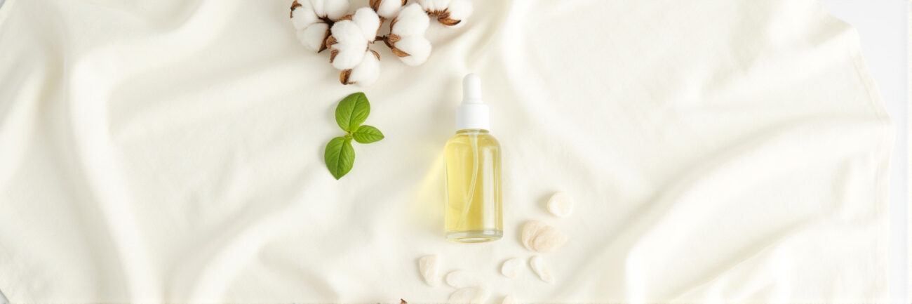 A clear glass perfume oil bottle on a white linen cloth, surrounded by cotton and green botanicals, representing 100% natural, clean, and alcohol-free formula luxury.